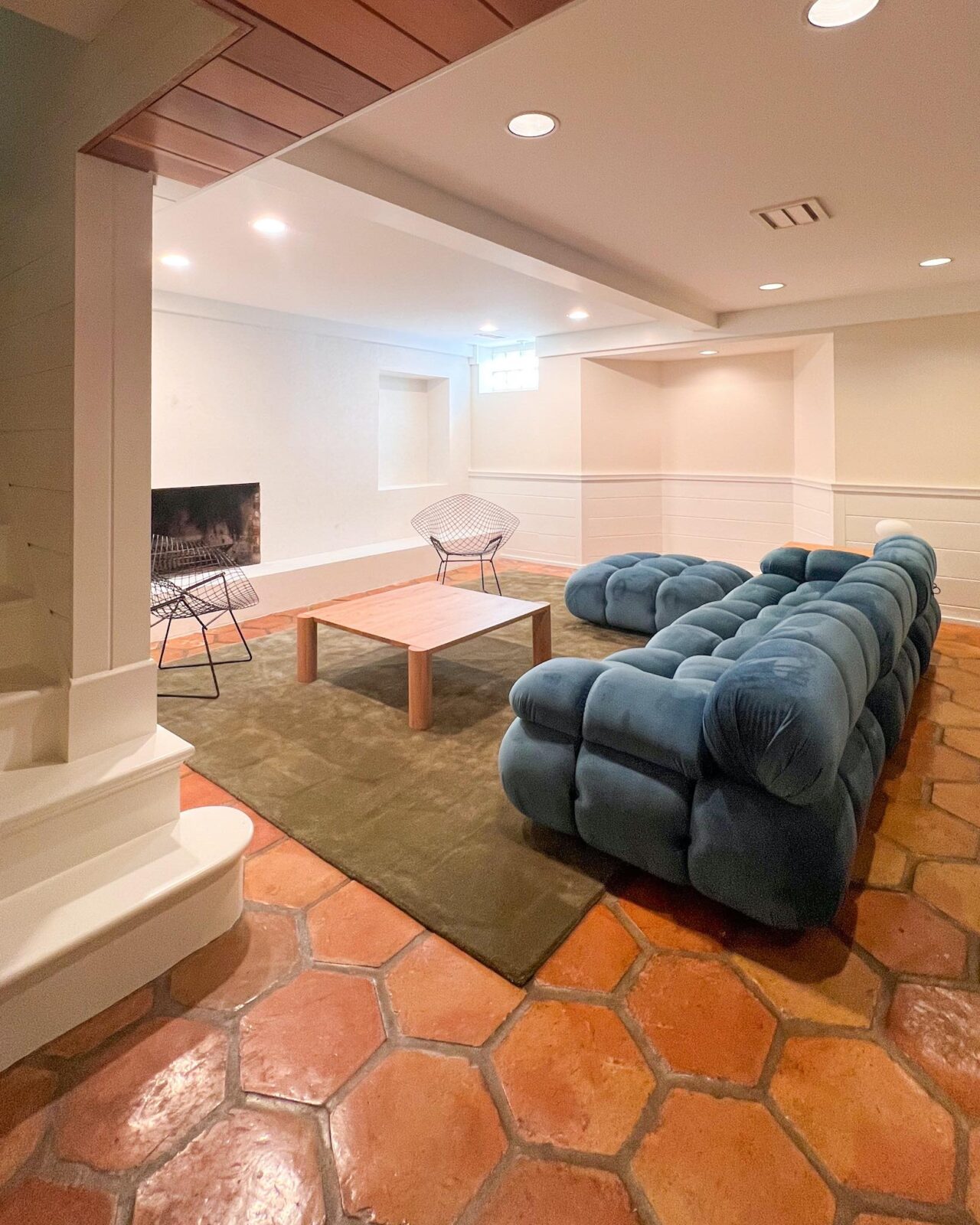 A basement family room with white and cream walls, plush green rugs, a blue modular sofa, a white oak coffee table, and vintage Bertoia diamond chairs