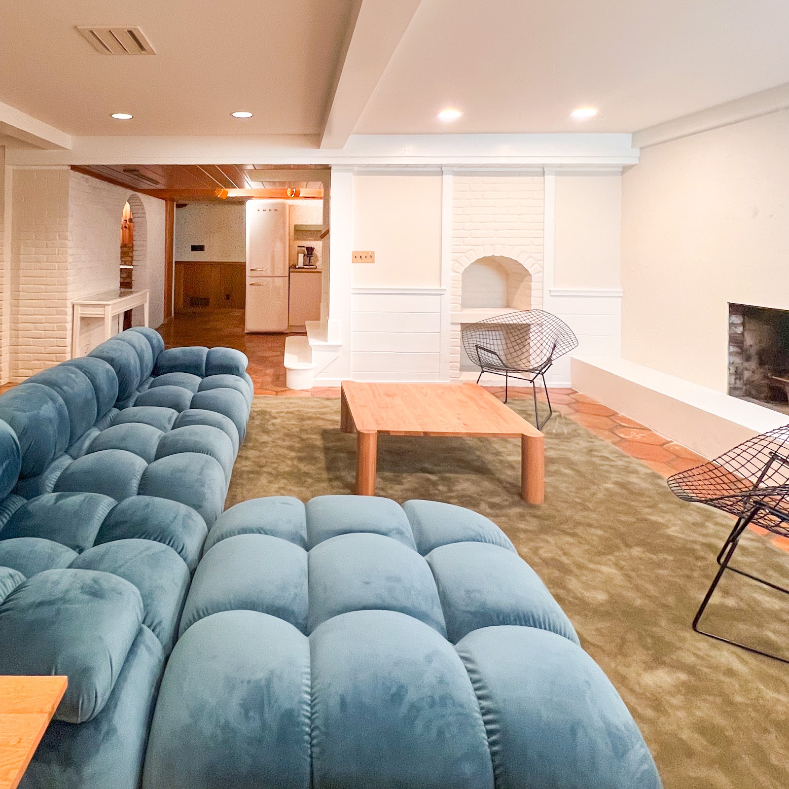 A basement family room with white and cream walls, plush green rugs, a blue modular sofa, a white oak coffee table, and vintage Bertoia diamond chairs