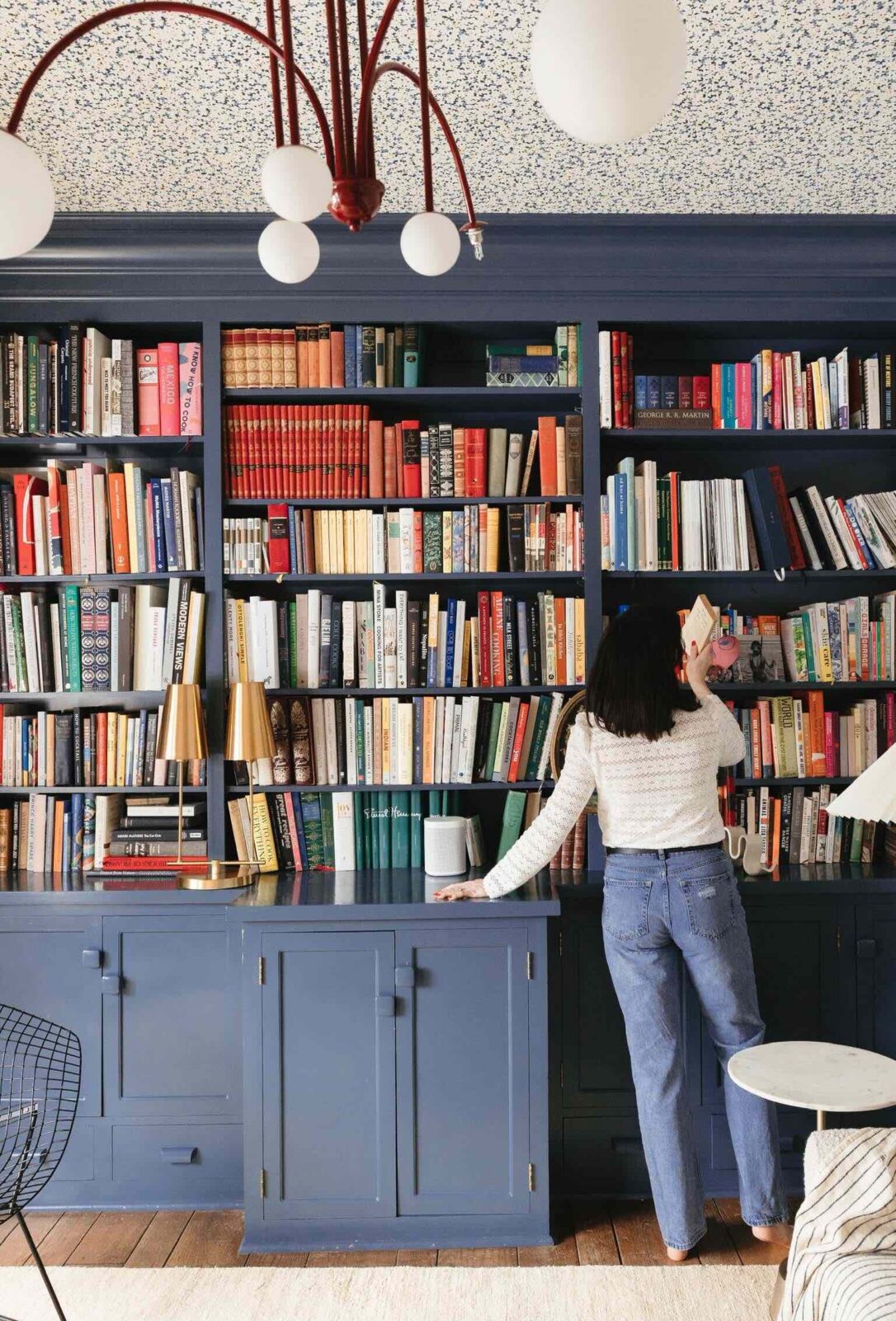 Woman is standing in front of a blue-painted built-in bookshelf, pulling a book off of a shelf.