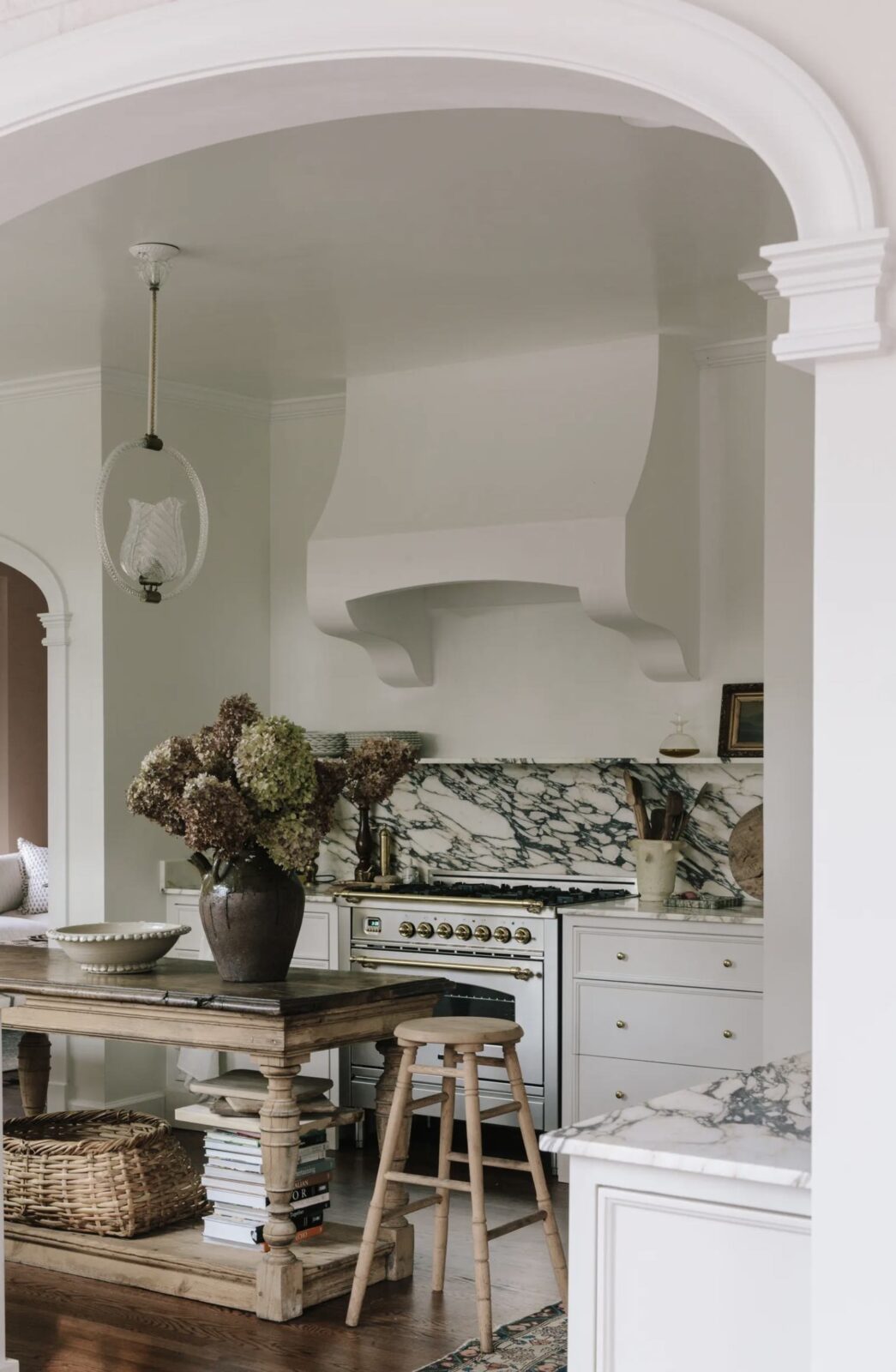 White kitchen with marble countertops, backsplash, and shelf, wood flooring, and antique wood island styled with wicker baskets, stacked cookbooks, and a large vase with dried hydrangeas