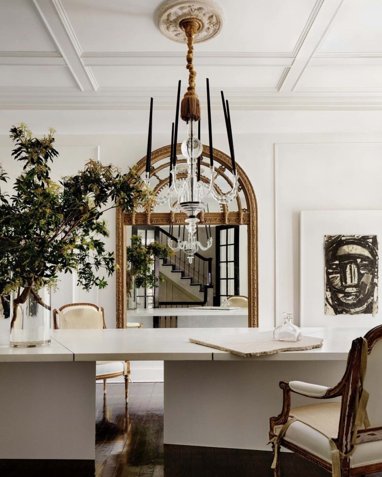 Dining room with white walls, large white dining table, antique gold upholstered chairs, chandelier, and large antique gilded mirror