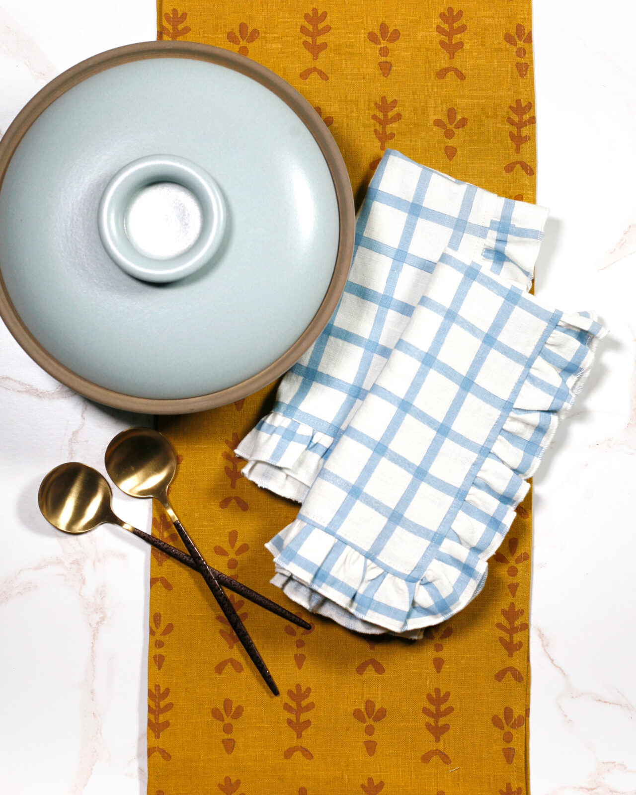 A yellow-printed table runner, blue and white dinner napkins, and serveware are displayed on a table.
