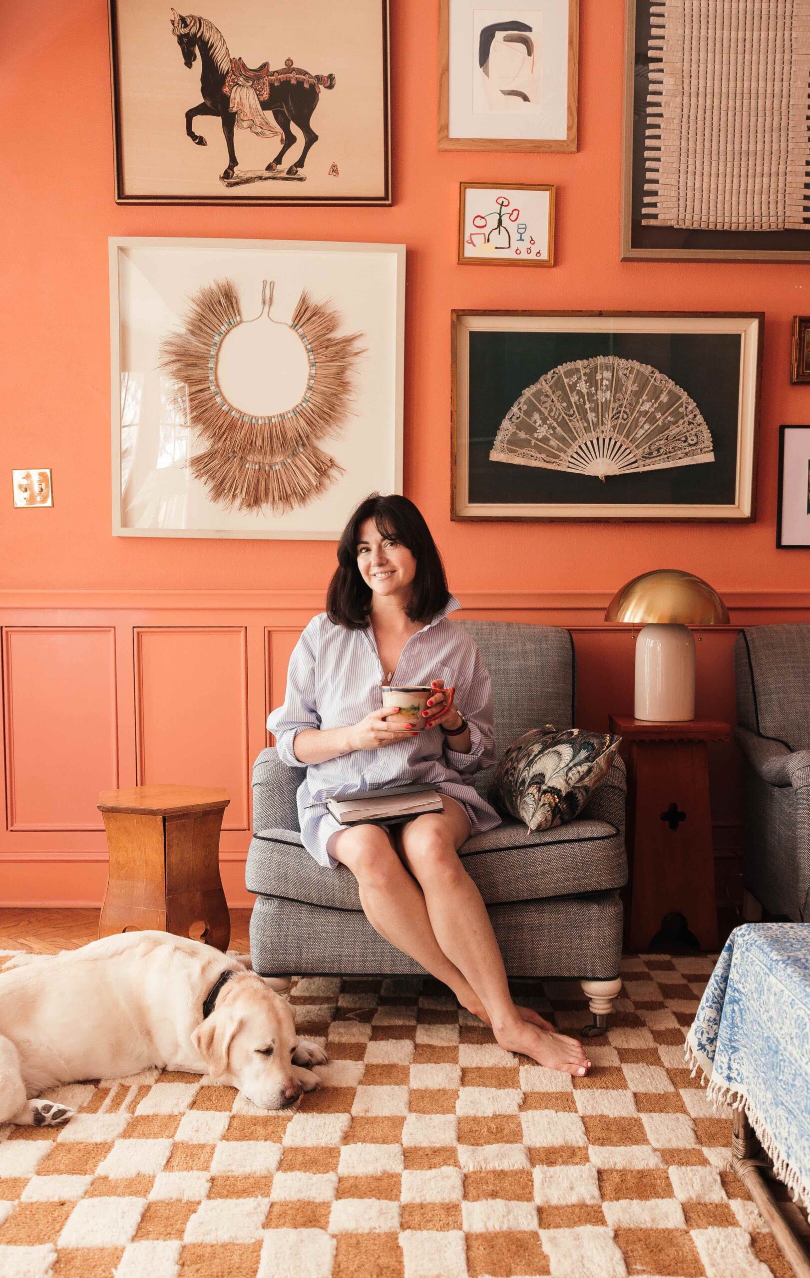 A woman holding a cup of coffee is sitting on a plush chair with a yellow lab sleeping at her feet. The wall behind her is painted a peach color and has a large gallery wall on display. The rug on the floor is in a brown and cream checkerboard pattern.