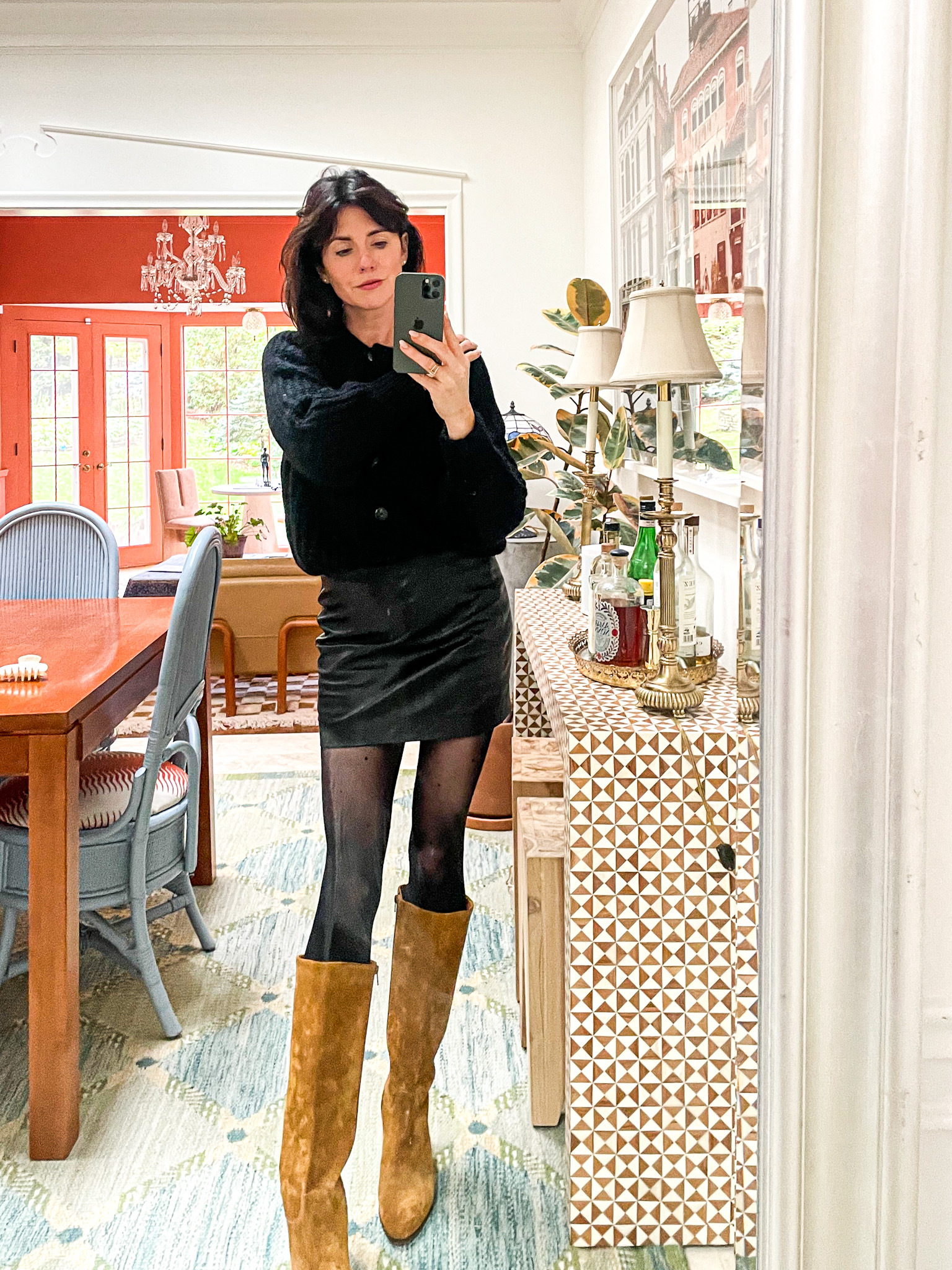 A woman is standing in front of a mirror wearing a black Sézane skirt, black cardigan sweater, black tights, and tall brown suede boots