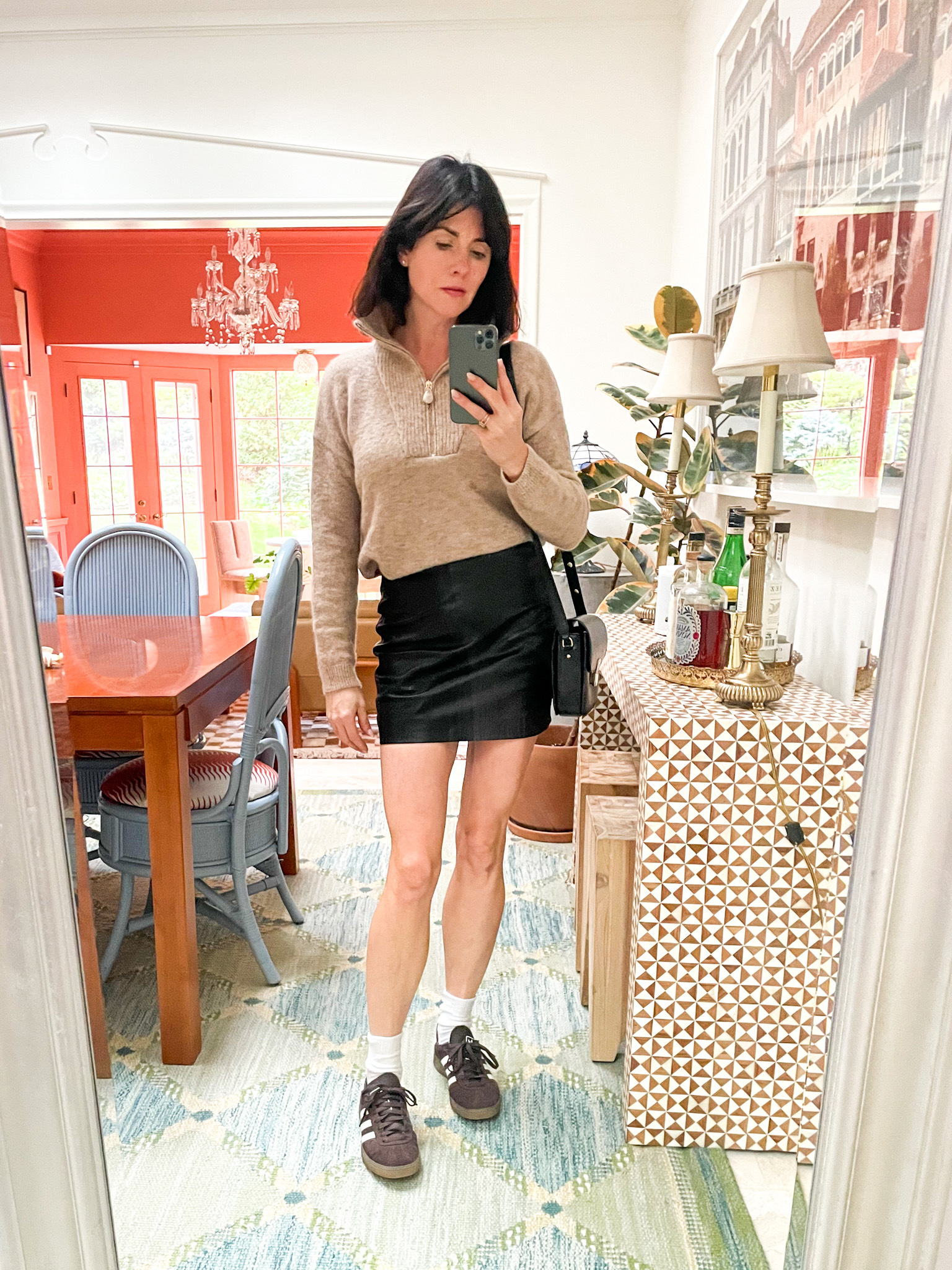 A woman is standing in front of a mirror wearing a black Sézane skirt, taupe half zip sweater, black crossbody bag, and brown adidas sneakers