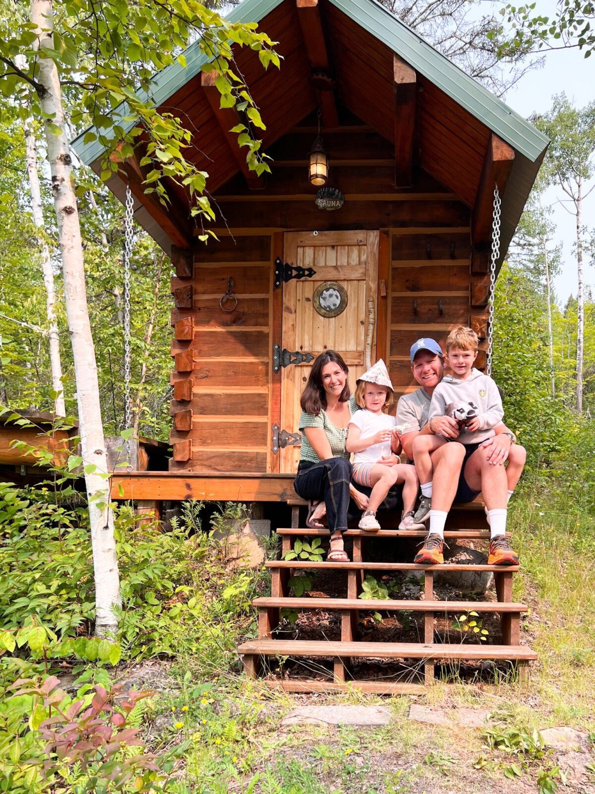 A family of four posing on the steps of a small log cabin in summer 2023