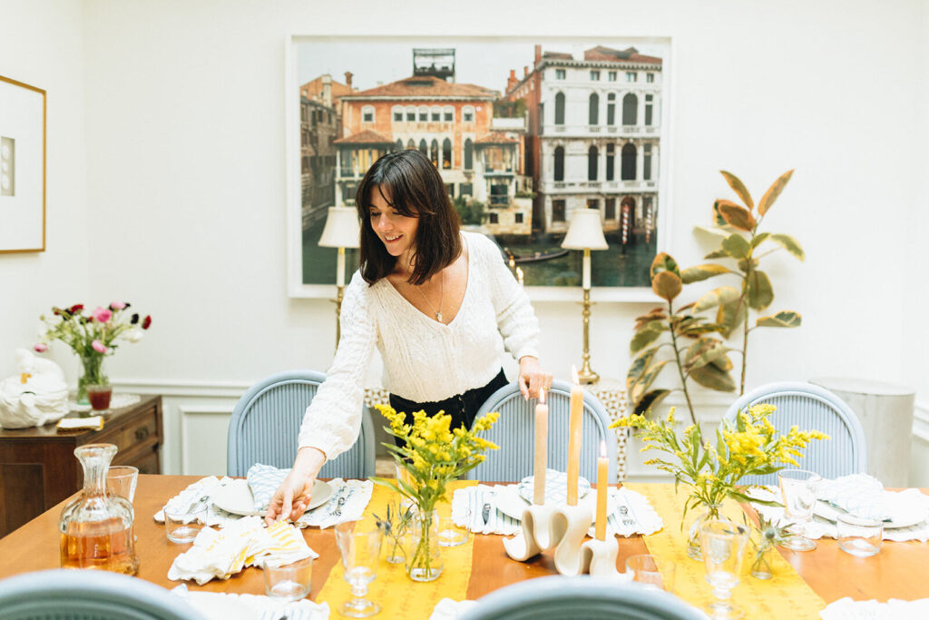 A woman is setting a wood dining table with tabletop linens, wine and water glasses, candles, and flower arrangements. A large printed photo of Venice, Italy is hung up on the wall behind her. 