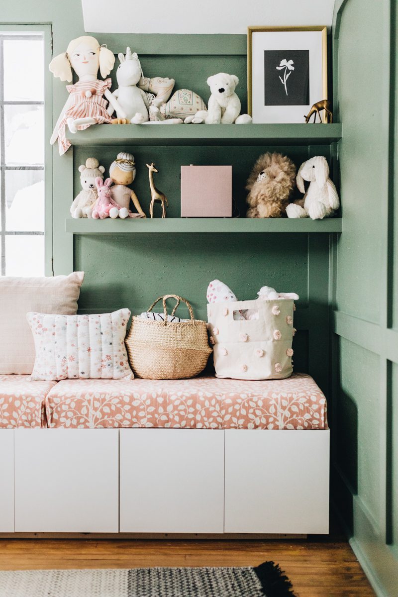 Nursery with green walls, a white bench with pink cushions, and lots of stuffed animals on shelves.